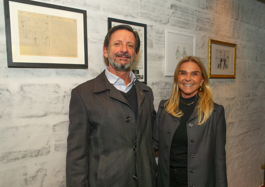André Jobim de Azevedo e Andréa Duarte, tendo ao fundo desenhos que contam etapas da trajetória de Vitório Gheno (Fotos de Evandro Leal/Divulgação)